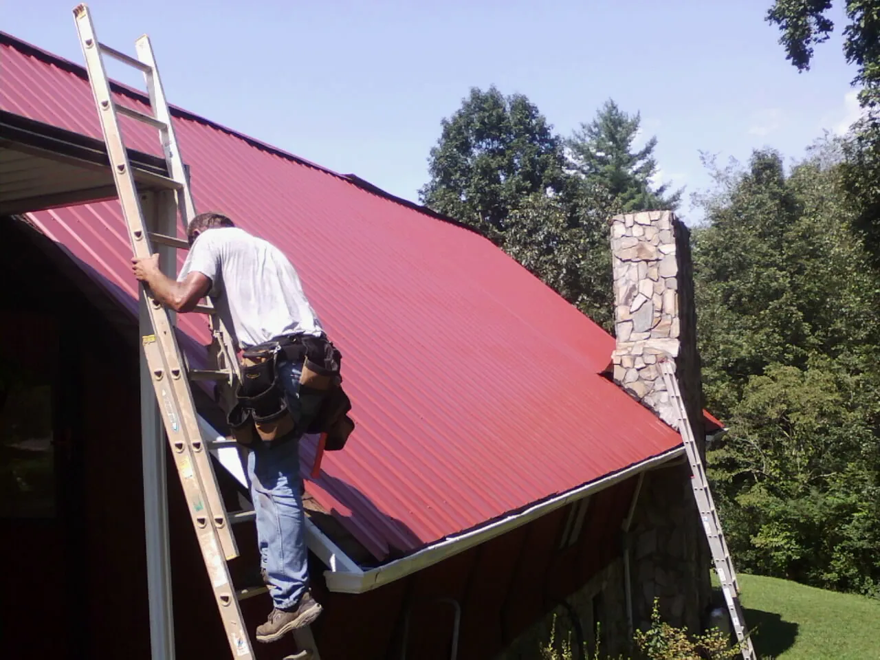 Roofer on a ladder beside a red metal roof and stone chimney — Soffit & Fascia Repair by Roofing Brothers in Loraine